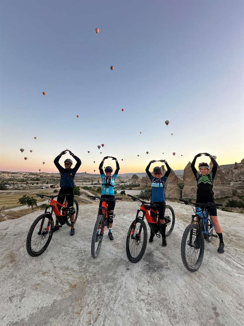 Vier fietsers steken de armen in de lucht onder de luchtballonnen bij zonsopgang